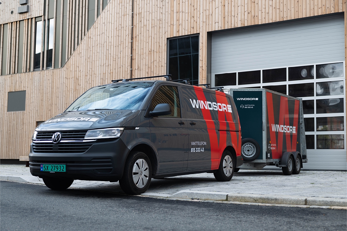 Mørk grå servicebil med tilhenger, dekorert med rød Windsor-logo, utenfor BIRs nybygg i Bergen